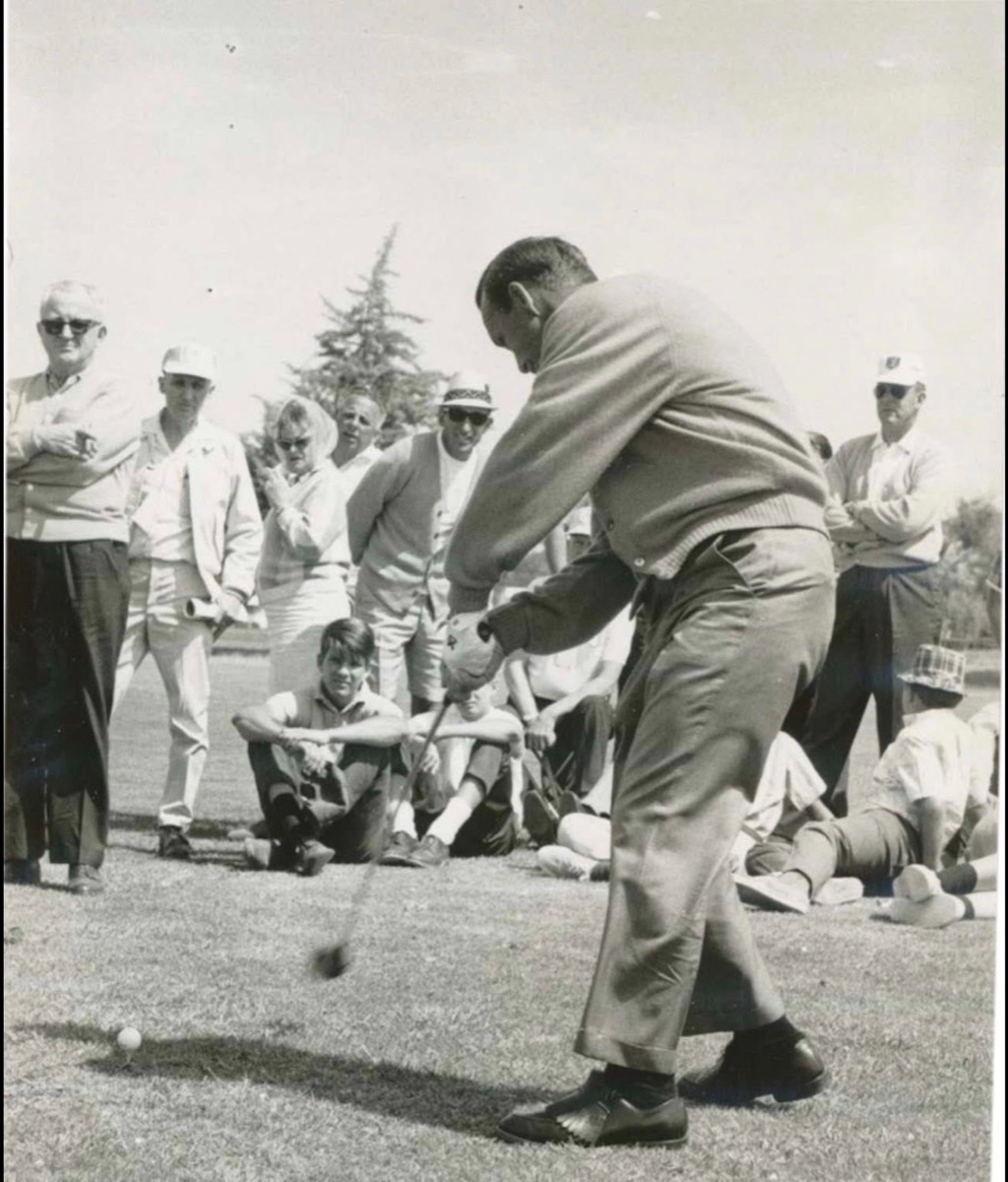 An archival black-and-white photograph of a golfer at impact in front of a gallery, his chest turned through the ball and his weight into the lead side